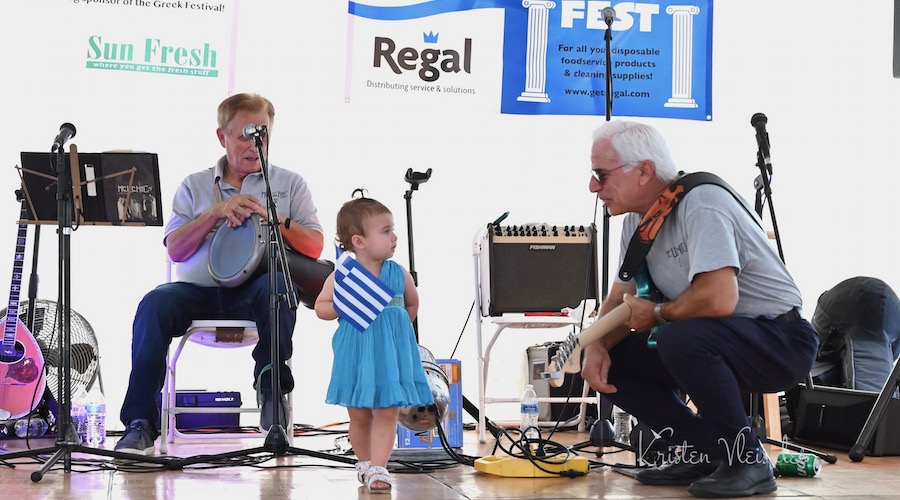 A toddler holds a Greek flag with two men onstage at Kansas City Greek Festival