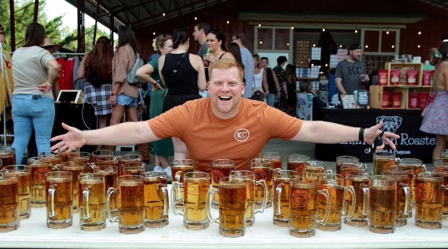 A man stands with his arms wide behind a table of full Oktoberfest steins